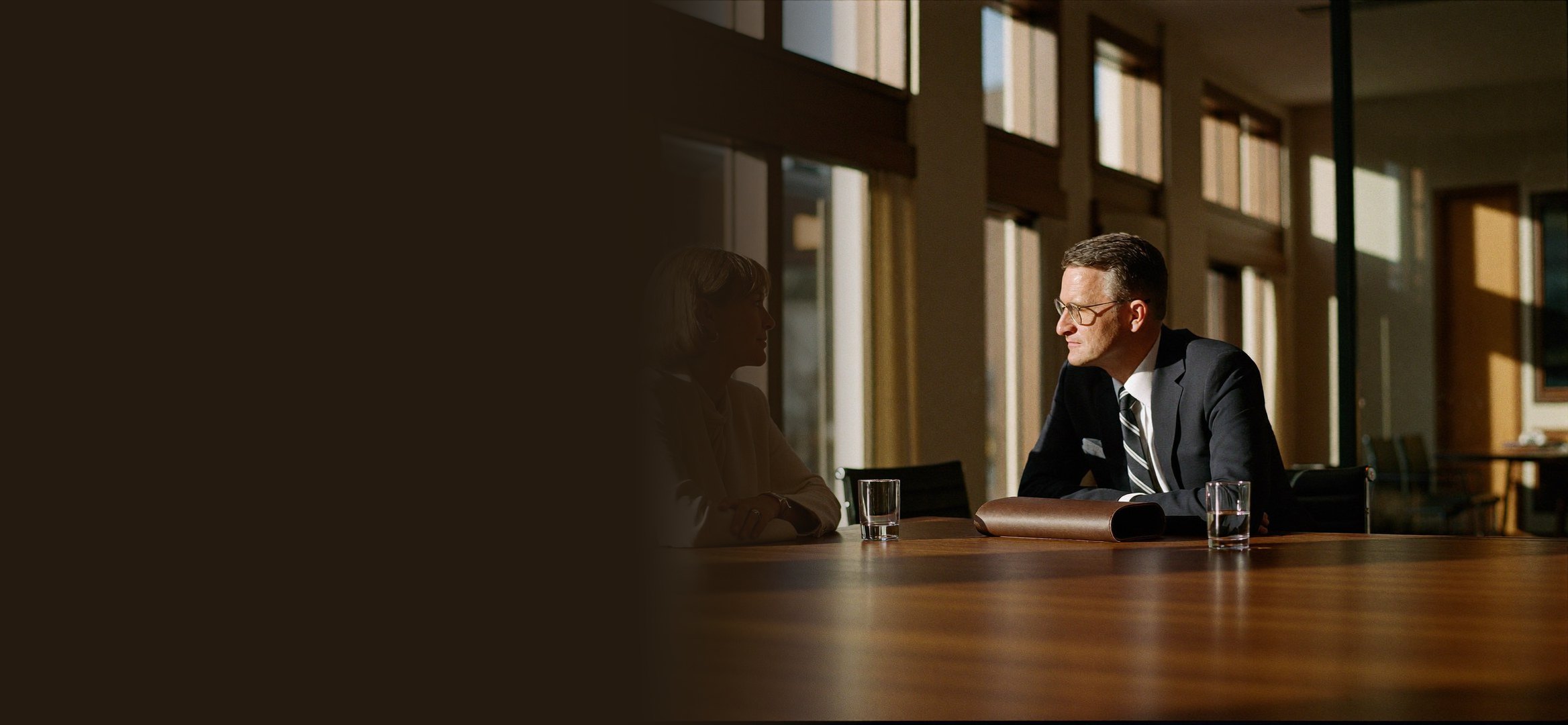 Two people in conversation at a long meeting table, afternoon light