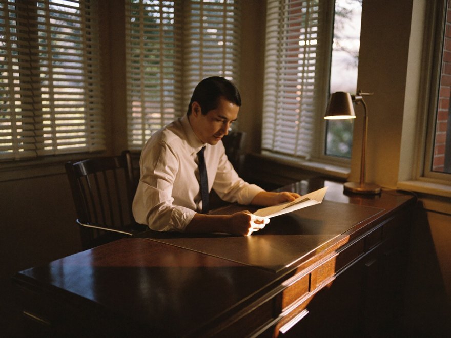 A business owner at a dark-wood desk by the window, reading, brass lamp lit