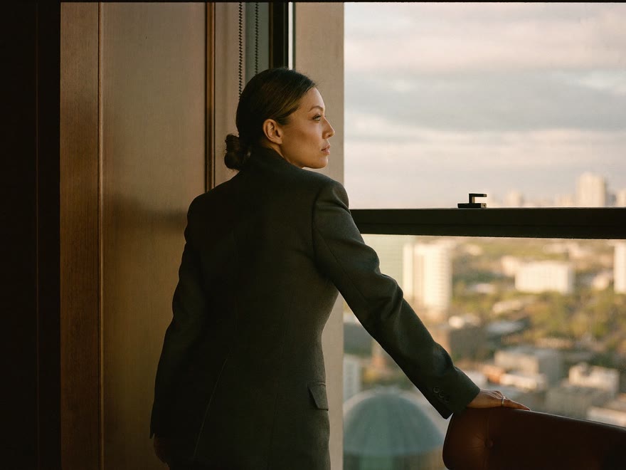 A business owner at the window of a panelled office, one hand on a chair back, looking out over the city
